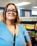 Angie, a woman wearing a blue shirt and glasses, stops for a smile in front of shelves of groceries.