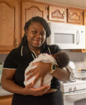 Woman Imani stands holding her infant child in the kitchen of her home. She smiles at the camera.