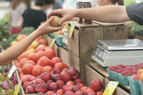 A hand reaches out, receiving an apple from a farmer's market vendor amidst other fresh produce