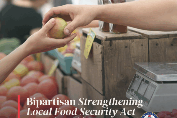 Graphic displays the RI Community Food Bank logo beside text box reading "Legislative Update." An image of an adult hand passing an apple to a child's hand at a farmer's market is featured. The header text reads "Bipartisan Strengthening Local Food Security Act Introduced in Senate"