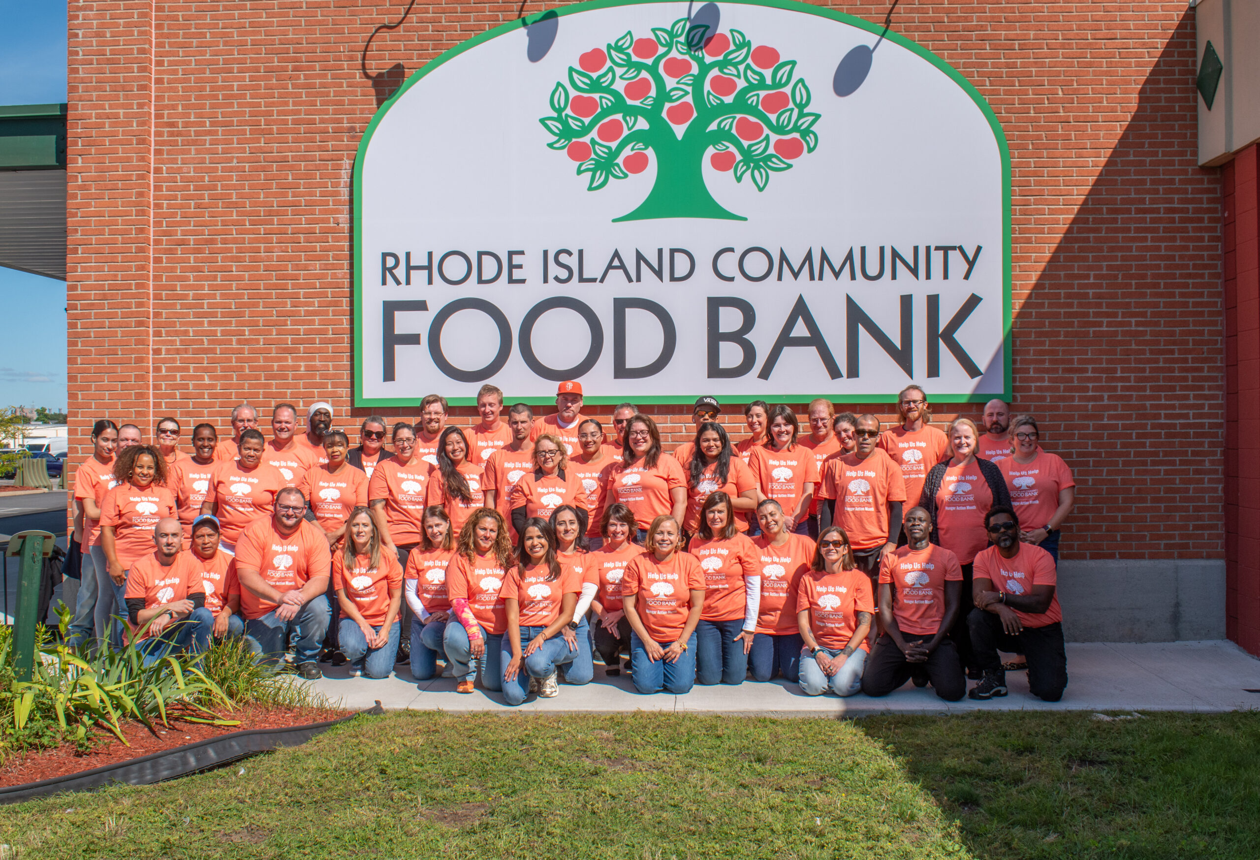 Rhode Island Community Food Bank staff all dress in matching orange Hunger Action Month t-shirts and group together for a photo in front of an outdoor wall with the RI Community Food Bank logo on it.